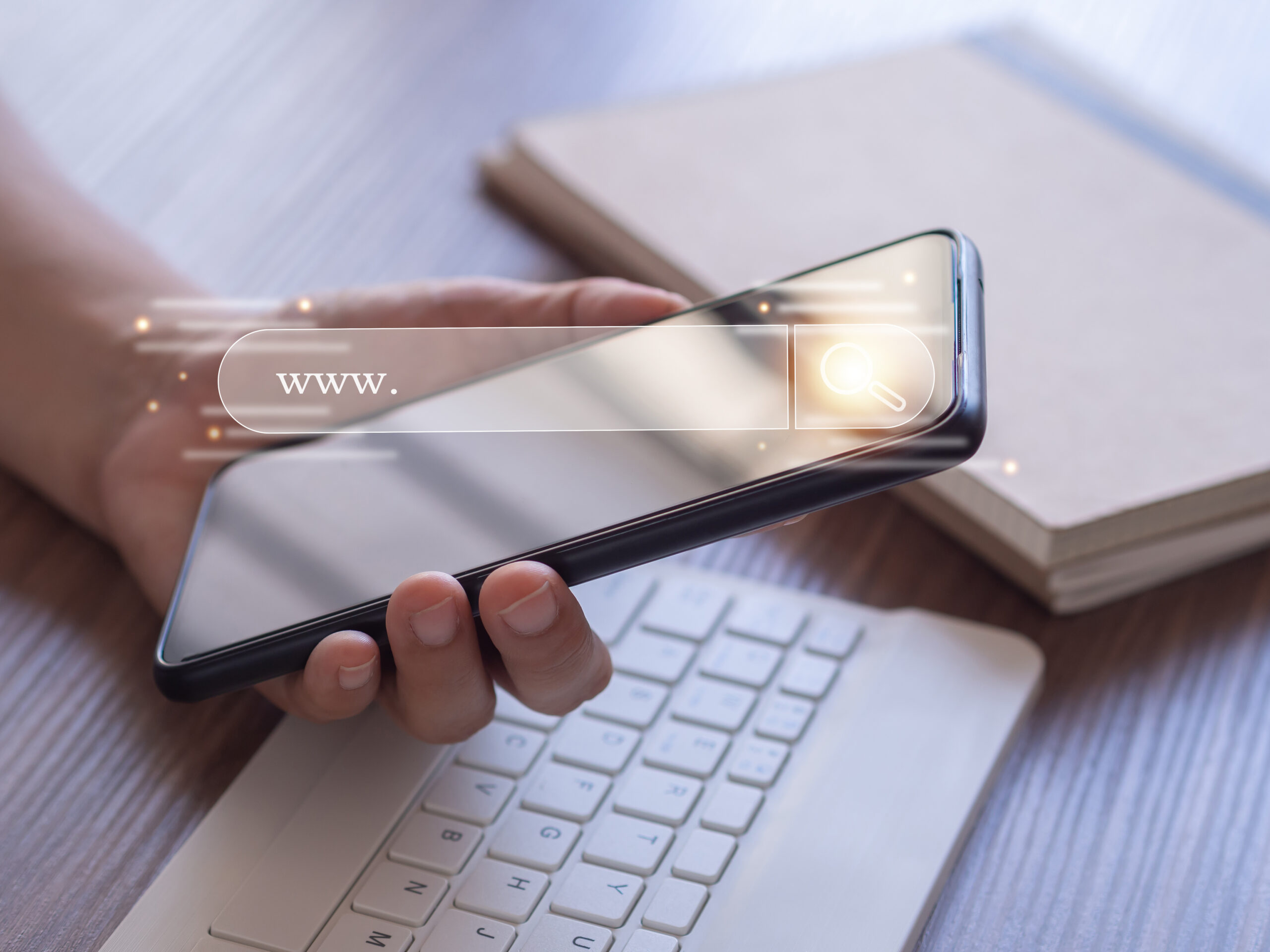 A hand holding a smartphone displaying a digital search bar, symbolizing beginner SEO concepts, placed near a keyboard and notebook on a wooden desk.