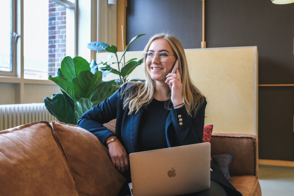 Smiling businesswoman talking on the phone with a laptop open, discussing a website redesign project in a modern office setting.